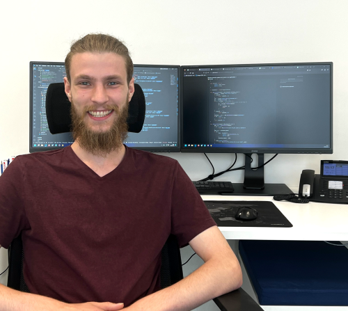 Smiling young employee at a workstation with two screens displaying code.